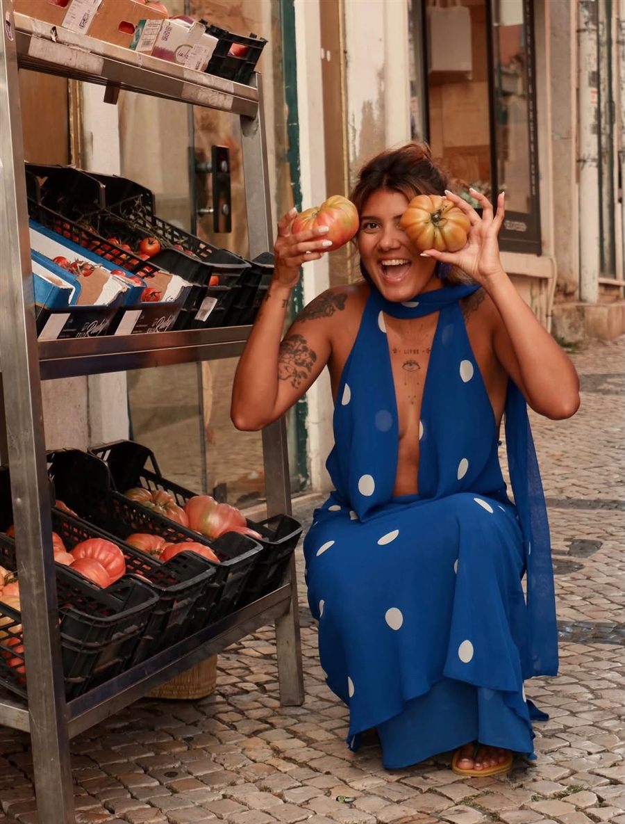 Thayna Auni agachada numa rua de Lisboa, sorrindo com dois tomates na mão